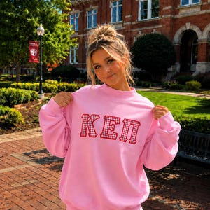 May include: A light pink long-sleeve sweatshirt featuring a mock neck collar and the letters KEP across the chest. The letters are rendered in a red and white gingham pattern with a thick red border, creating a bold, textured graphic design.