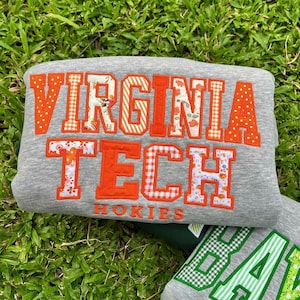 May include: A heather gray sweatshirt featuring the text VIRGINIA TECH HOKIES in large, block-style letters. The letters are appliqued with a mix of solid orange fabric and various patterns, including orange polka dots, floral prints, and gingham.