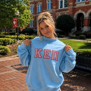 May include: A light blue crewneck sweatshirt featuring the letters KEP in a red and white gingham pattern. The sweatshirt has long sleeves, a ribbed mock neck collar, and a relaxed fit, with the letters appliqued across the center of the chest.