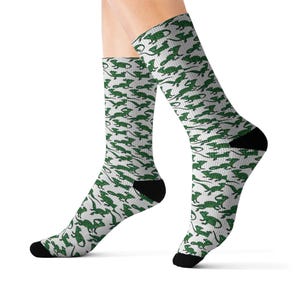 May include: A pair of crew socks with a repeating pattern of green lizards on a white background. The socks have black heels and toes. The socks are made of a soft, comfortable material.