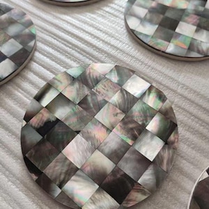 May include: A collection of round coasters with a checkered pattern made from iridescent mother-of-pearl. The coasters have a neutral color palette with hints of green, pink, and gray. The coasters are arranged on a white surface.