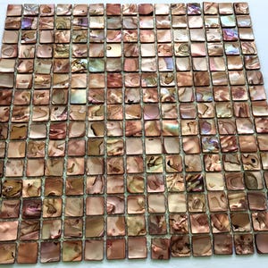 Może przedstawiać: Kwadratowy arkusz płytek mozaikowych składający się z wielu małych, iryzujących, kwadratowych płytek. Płytki prezentują gamę ciepłych, ziemistych odcieni, w tym brązy, beże i nuty różu i fioletu. Powierzchnia ma odblaskową, perłową jakość.