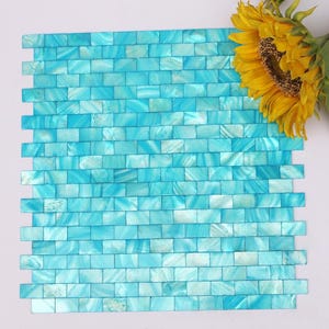 May include: A square of iridescent blue mosaic tiles in a brick pattern. The tiles have a pearlescent sheen, with variations of light and dark blue. A yellow sunflower with a brown centre is in the upper right corner.