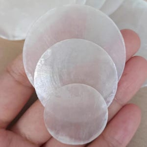 May include: Several translucent, circular mother-of-pearl discs in different sizes. The pearly white surfaces have an iridescent quality, reflecting light. The discs are held in a hand, indicating their potential use in crafts or jewellery making.