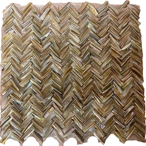 Pode incluir: Uma folha de azulejo de mosaico com um padrão de espinha de peixe. Os azulejos são uma mistura de cores douradas iridescentes, castanhas e creme. Os azulejos têm uma aparência natural e texturizada e estão dispostos em forma de V repetida.