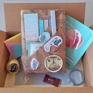 Może przedstawiać: Pudełko prezentowe wypełnione różnymi przedmiotami, w tym maską do twarzy, kulą do kąpieli, gumką do włosów, naklejkami i zapakowanym pakietem. Pudełko jest brązowe, a przedmioty są ułożone w środku. Zapakowany pakiet ma brązowy wzór liści.