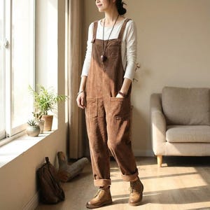 May include: Brown corduroy overalls with large front pockets and adjustable straps. The overalls are worn with a white long-sleeved top and brown boots. The outfit is casual and comfortable, suitable for everyday wear.