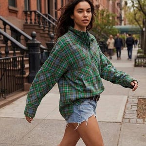 Könnte beinhalten: Ein langärmliges Hemd mit grünem, blauem und gelbem Karomuster und einem Kragen mit halbem Reißverschluss. Das Hemd wird mit Jeansshorts kombiniert. Die Person geht auf einem Bürgersteig vor einem Backsteingebäude.