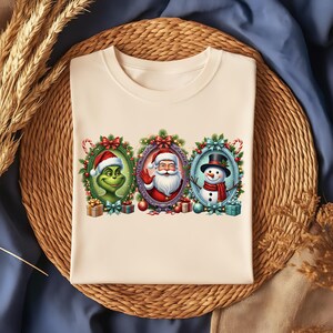 Puede incluir: Camiseta color crema con tres personajes navideños: El Grinch, Papá Noel y un muñeco de nieve, cada uno enmarcado en un óvalo con decoraciones festivas. El Grinch lleva un gorro de Papá Noel, Papá Noel saluda y el muñeco de nieve tiene un sombrero de copa y una bufanda.