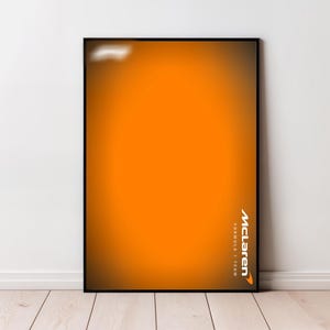 Puede incluir: Póster enmarcado con un fondo degradado naranja. El logotipo de McLaren Formula 1 Team se muestra en blanco y naranja en la esquina inferior derecha. El póster está colocado contra una pared blanca y un suelo de madera clara.