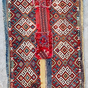 May include: A rectangular, hand-woven textile with a geometric pattern in red, blue, white, and brown. The design features repeating diamond shapes. A central red panel and striped borders add to the visual interest. The textile appears to be aged.