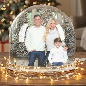 May include: A clear glass snow globe with a family portrait inside, illuminated by a string of warm white lights. The globe sits on a wooden surface, decorated with Christmas ornaments and greenery. A Christmas tree is in the background.