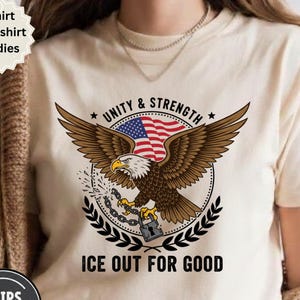 Puede incluir: Camiseta color crema con un gráfico detallado de un águila calva americana con las alas extendidas, sosteniendo una cadena y un candado. El águila tiene un diseño patriótico de la bandera estadounidense en el pecho. El texto "UNITY & STRENGTH" y "ICE OUT FOR GOOD".