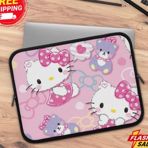 May include: Pink laptop sleeve featuring a repeating pattern of Hello Kitty characters and teddy bears. The sleeve has a black zipper and trim. The design includes bows, hearts, and speech bubbles on a pink background. The sleeve is on a wooden surface.