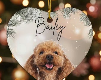 Puede incluir: Adorno navideño en forma de corazón con un perro marrón sonriente llamado Bailey. El adorno cuelga frente a un árbol de Navidad decorado. El fondo está borroso con luces y adornos navideños. El adorno tiene una cuerda dorada para colgar.