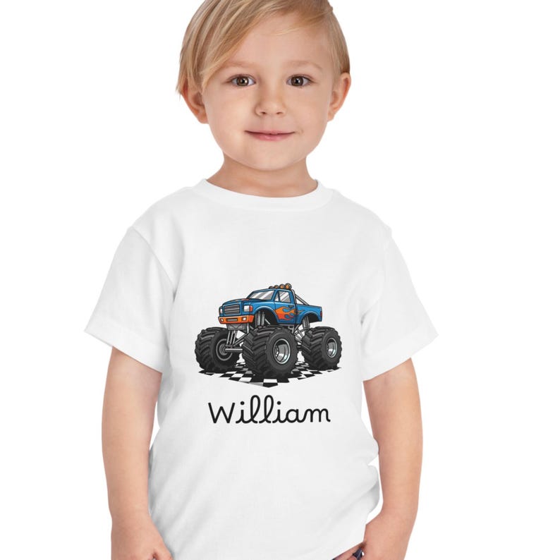 May include: White toddler t-shirt featuring a blue monster truck graphic with orange and yellow flames. The truck has large black tires and is set on a checkered pattern. The name "William" is printed below the truck in black.