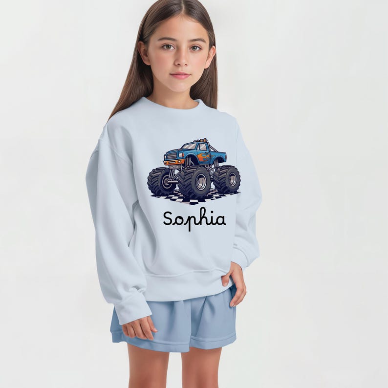 May include: Light blue sweatshirt with a graphic of a blue monster truck with flames and large tires. The name "Sophia" is printed in black script below the truck. The sweatshirt is paired with light blue shorts.