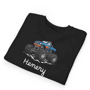 May include: Black t-shirt featuring a blue monster truck with orange flames and large black tires. The truck is on a checkered flag design. The name "Henery" is written in white script below the truck.
