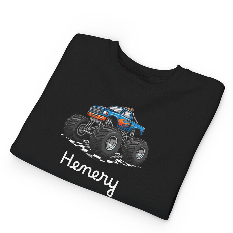 May include: Black t-shirt featuring a blue monster truck with orange flames and large black tires. The truck is on a checkered flag design. The name "Henery" is written in white script below the truck.