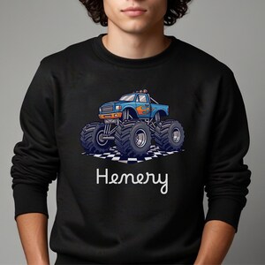 May include: Black sweatshirt featuring a blue monster truck graphic with orange flames and large tires. The name "Henery" is written in white script below the truck. The truck is on a checkered pattern.