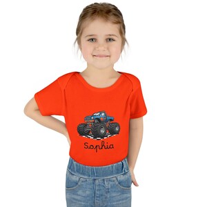 May include: An orange baby bodysuit featuring a blue monster truck graphic with flames and large tires. The name "Sophia" is written in cursive below the truck. The bodysuit is short-sleeved and made of a soft material.