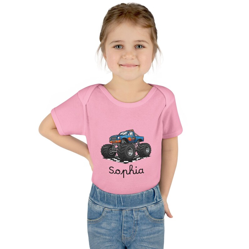 May include: Pink short-sleeve bodysuit with a blue monster truck graphic and the name "Sophia" in cursive. The truck has large black tires and is set against a checkered background.