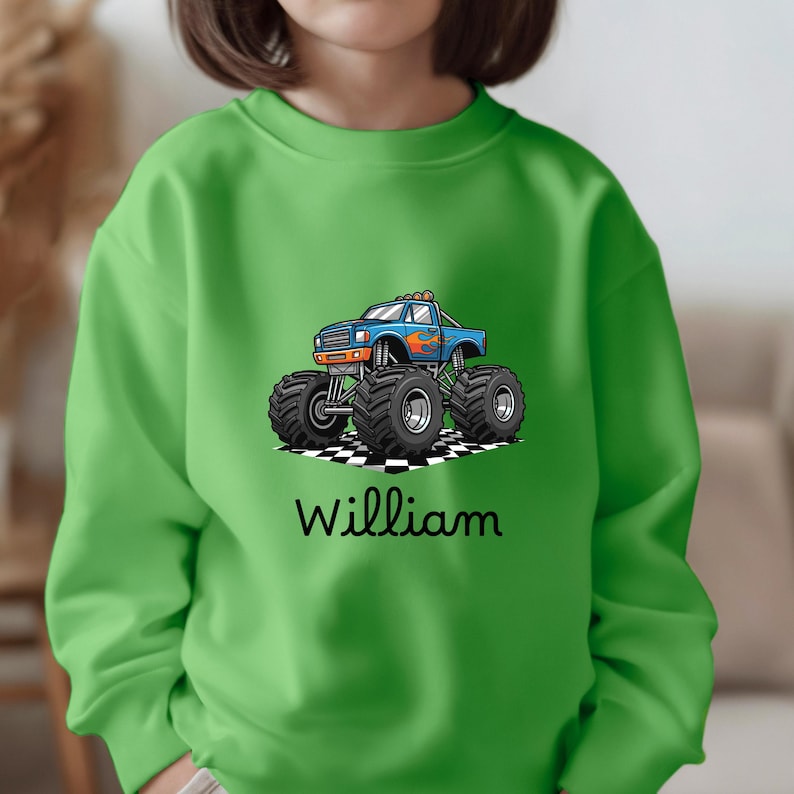 May include: Green sweatshirt featuring a blue monster truck graphic with orange and red flames. The truck is on a checkered pattern, and the name "William" is printed below in black script.