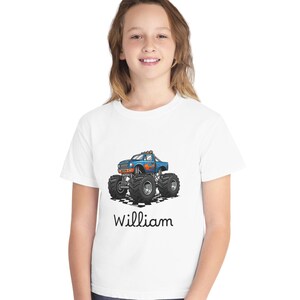 May include: White t-shirt featuring a blue monster truck graphic with orange flames and large black tires. The name "William" is printed in black script below the truck. The shirt is worn by a person.