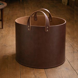 May include: A cylindrical, brown leather log carrier with two arched handles. The handles are attached to the body with brass rivets. The interior of the carrier is a lighter shade of brown. The carrier is sitting on a wooden floor.