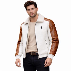 Puede incluir: Chaqueta blanca con mangas de cuero sintético marrón y cuello de borreguillo color crema. La chaqueta presenta un logotipo negro en el pecho y cierre de cremallera. Una prenda de abrigo elegante.