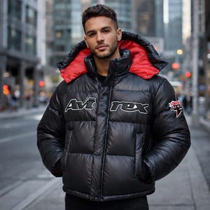 May include: Black puffer jacket with a red hood lining. The jacket features the white text "Avi Rex" across the chest and a patch on the sleeve. The jacket is worn in an urban setting.