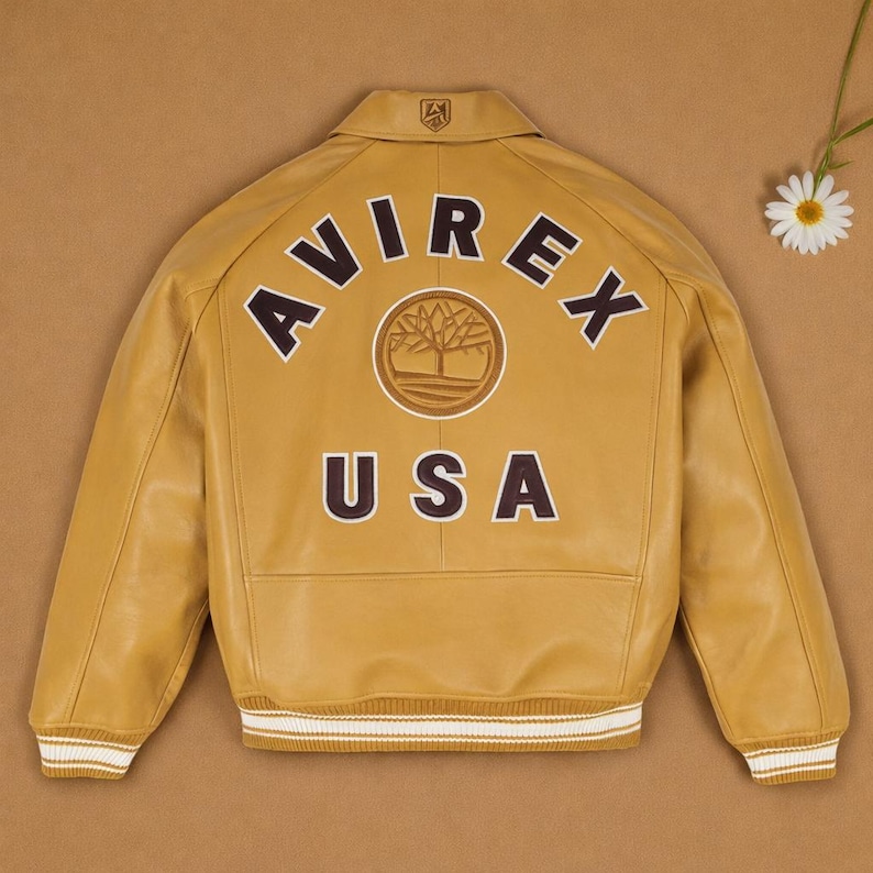 May include: Mustard yellow leather jacket with the word "AVIREX" arched across the back in dark brown lettering. Below the word "AVIREX" is a circular emblem with a tree design. The word "USA" is printed below the emblem. The jacket has white and cream striped cuffs.