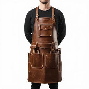 May include: A brown leather apron with multiple pockets, straps, and rivets. The apron has a chest pocket with a flap and a zippered pocket. Tools, including a hammer and pliers, are visible in the pockets. The apron is designed for craftspeople.