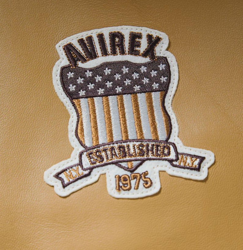 May include: Embroidered patch with the text "AVIREX ESTABLISHED NY 1975" in brown and gold. The patch features a shield design with the American flag and star accents, set against a tan background.