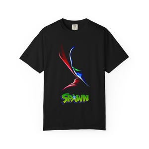 Puede incluir: Una camiseta negra de manga corta con un gráfico del personaje Spawn. El diseño muestra una silueta estilizada con reflejos rojos, azules y verdes. Debajo del personaje, la palabra "SPAWN" está impresa en una fuente verde y negrita.