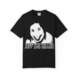 Puede incluir: Una camiseta negra de manga corta con un gráfico blanco de alto contraste que muestra un rostro de ojos muy abiertos y sonrisa amplia con cabello oscuro largo. Debajo, el texto JEFF THE KILLER está impreso en una fuente blanca estilizada y goteante.