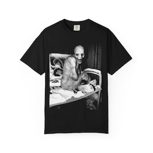 May include: A black short-sleeve t-shirt featuring a high-contrast black and white graphic print on the chest. The image depicts a pale, gaunt figure with large eyes and a wide, toothy grin sitting on a bed frame. The shirt has a crew neckline.