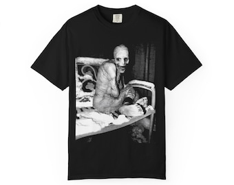 May include: A black short-sleeve t-shirt featuring a high-contrast black and white graphic print on the chest. The image depicts a pale, gaunt figure with large eyes and a wide, toothy grin sitting on a bed frame. The shirt has a crew neckline.