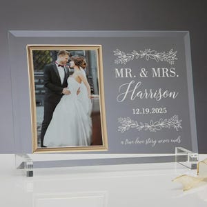 May include: A rectangular glass photo frame with a gold-toned border containing a photograph of a couple in wedding attire. The glass is etched with the text MR. & MRS. Harrison, the date 12.19.2025, floral accents, and the phrase a true love story never ends.