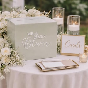 May include: A sage green wedding card box with "Mr. & Mrs. Oliver" in white script. A gold-framed sign reads "Cards". The table is decorated with white flowers, candles in glass holders, and a gold tray with stationery.