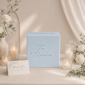 May include: A light blue rectangular card box with a slot on top, featuring the raised white script text The Nicholsons on the front. A small white card with the text Thank You for celebrating with us is placed in front of the box.