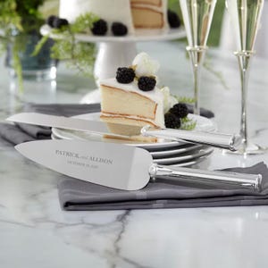 May include: A silver-toned cake knife and server set with polished handles. The server blade is engraved with the text PATRICK and ALLISON OCTOBER 19, 2026. The utensils are positioned on a grey cloth napkin in front of a slice of cake.