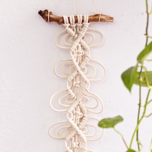 May include: A white macrame wall hanging with a natural wood branch at the top. The macrame is made with a knotting technique that creates a series of loops and swirls.