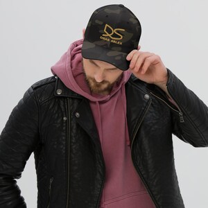 May include: A black and brown camouflage baseball cap with the gold text "DRAS SELEX" and a stylized "DS" logo. The cap is worn by a person wearing a pink hoodie and black leather jacket.