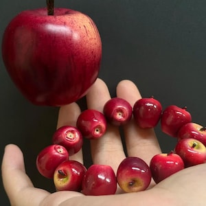 Puede incluir: Una gran manzana roja con un tallo oscuro flota sobre una mano que sostiene un anillo de pequeñas manzanas rojas. Las manzanas tienen un acabado brillante y una apariencia realista, lo que sugiere un producto con temática de frutas.