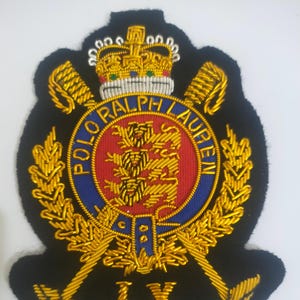 Könnte beinhalten: Gestickter Aufnäher mit Krone, gekreuzten Rudern und dem Text "POLO RALPH LAUREN". Der Aufnäher hat einen schwarzen Hintergrund mit goldenen und roten Akzenten. Die römischen Ziffern "LX" befinden sich unten.