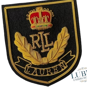 Könnte beinhalten: Schwarzer, schildförmiger, gestickter Aufnäher mit goldener Umrandung. Verfügt über eine rote und goldene Krone über den goldenen Buchstaben "RL". Unter den Buchstaben befinden sich goldene Blattmotive und das Wort "LAUREN" in Gold.