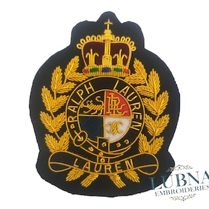 Könnte beinhalten: Ein schwarzer, gestickter Aufnäher mit goldenen und roten Akzenten. Der Aufnäher zeigt eine Krone, Lorbeerblätter und die Worte "RALPH LAUREN" in einem kreisförmigen Design. Die Mitte hat ein Schilddesign mit mehreren Farben.