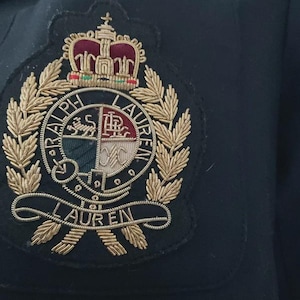 Könnte beinhalten: Nahaufnahme eines marineblauen Blazers mit einem gestickten Wappen. Das Wappen zeigt eine Krone, Lorbeerblätter und den Namen "RALPH LAUREN" in Goldfaden. Die Tasche des Blazers ist sichtbar.