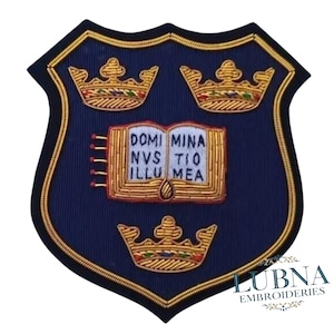 May include: A navy blue embroidered shield-shaped patch with gold detailing. It features three gold crowns and an open book with the Latin inscription "DOMI MINA NVS TIO ILLU MEA." The border is gold and black.